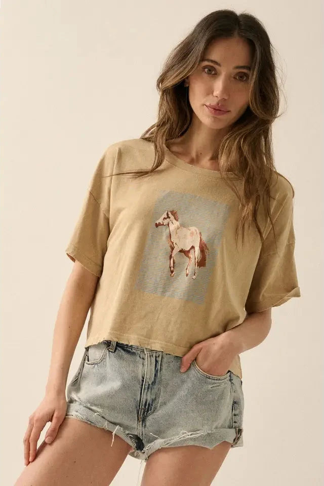 A woman wearing a beige graphic t-shirt with a vintage-style horse graphic print and striped background, round neckline, short sleeves, distressed detailing at neckline, cuffs, and hem, paired with denim shorts.