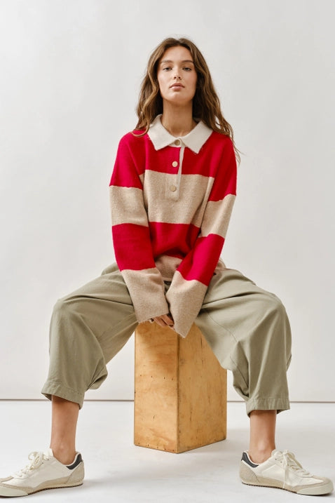 Woman wearing a red and beige striped sweater with a white collar, sitting on a wooden block against a plain background.