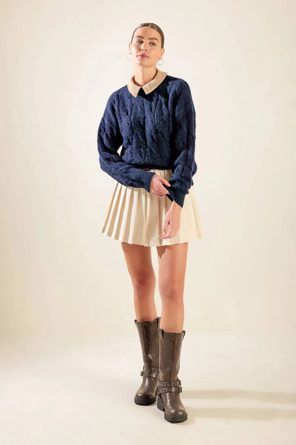 Person wearing a blue cable knit sweater, beige pleated skirt, and brown boots on a plain background