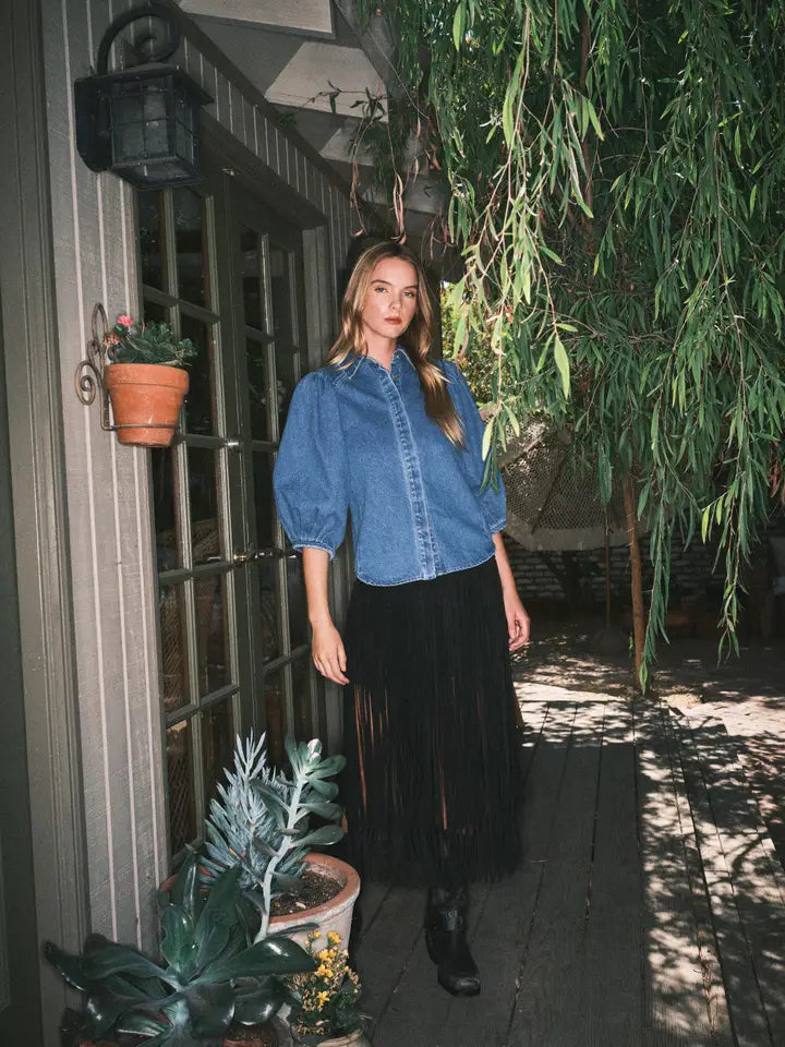 Woman wearing a blue denim shirt and black skirt standing outdoors with plants around.
