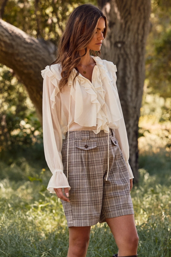 Woman wearing a white blouse and plaid shorts standing in a natural setting with trees and grass.