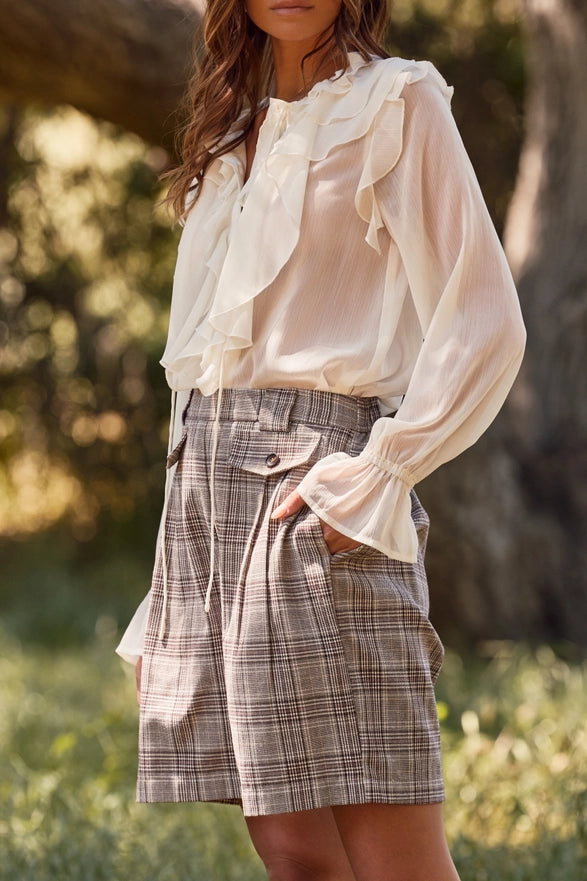 Woman wearing a white blouse with ruffled sleeves and plaid shorts in an outdoor setting.