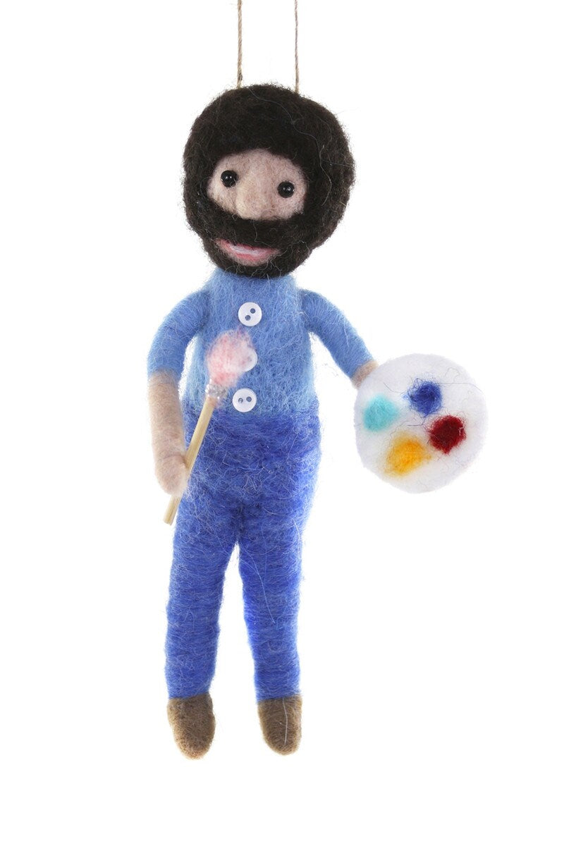 Felted ornament of a Bob Ross wearing a blue outfit with a paintbrush and palette on a white background