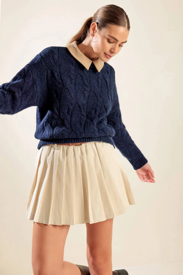 Woman wearing a navy blue cable knit sweater and cream pleated skirt on a beige background
