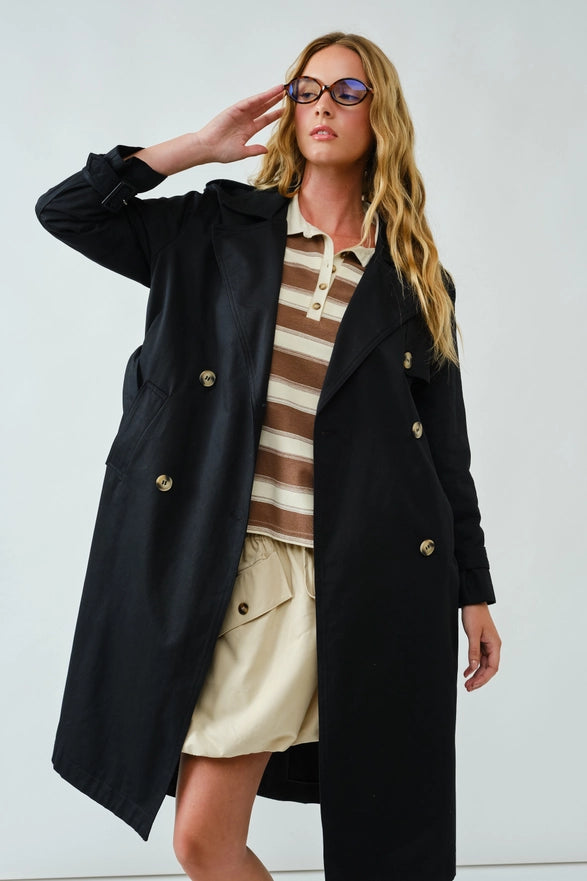 Woman wearing a black coat with gold buttons, striped shirt, and beige skirt on a white background
