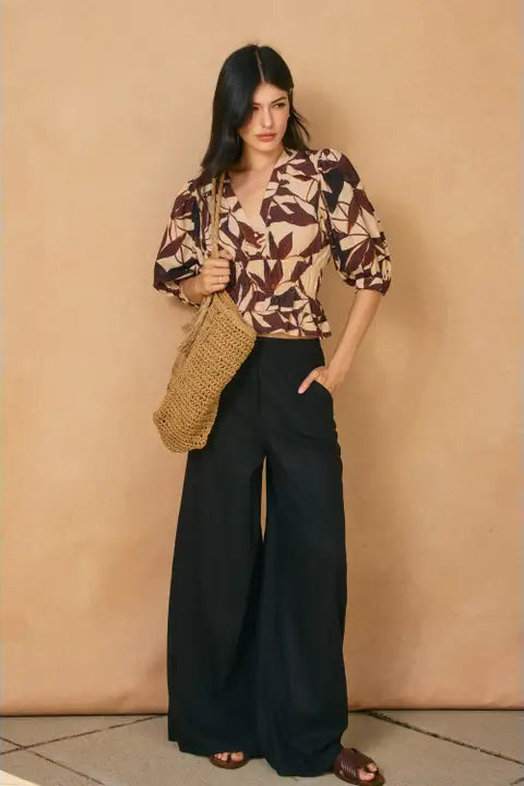 Woman wearing a patterned top and black pants, holding a woven bag against a beige wall.