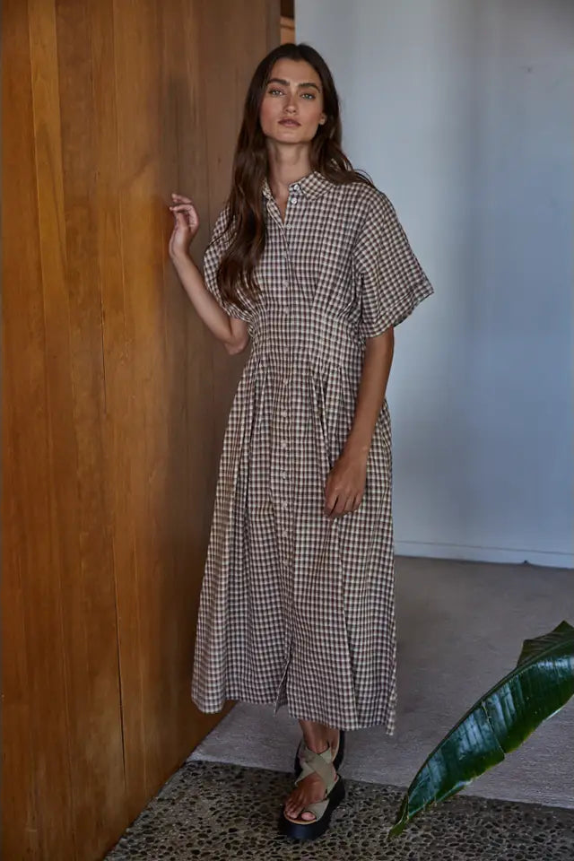 Woman wearing a checkered dress standing in a room with wooden walls and a plant.