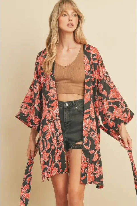 A woman wearing a floral midi kimono with a sash tie belt and side slits, paired with a brown top and denim shorts.