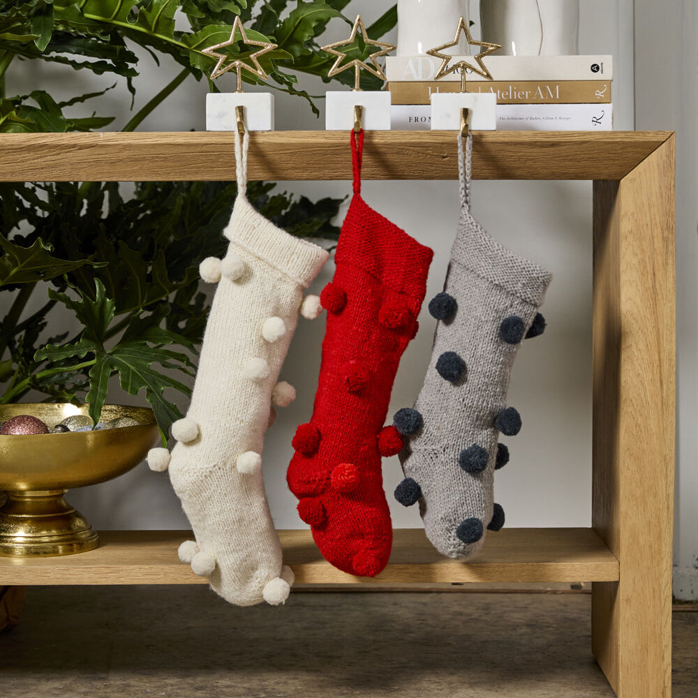 Three Christmas stockings in white, red, and gray hanging on a wooden shelf with plants and decor in the background.