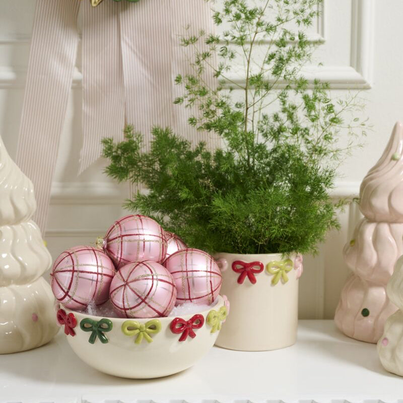 Decorative scene with a wreath, bowls of pink and gold ornaments, and a potted plant on a white surface.