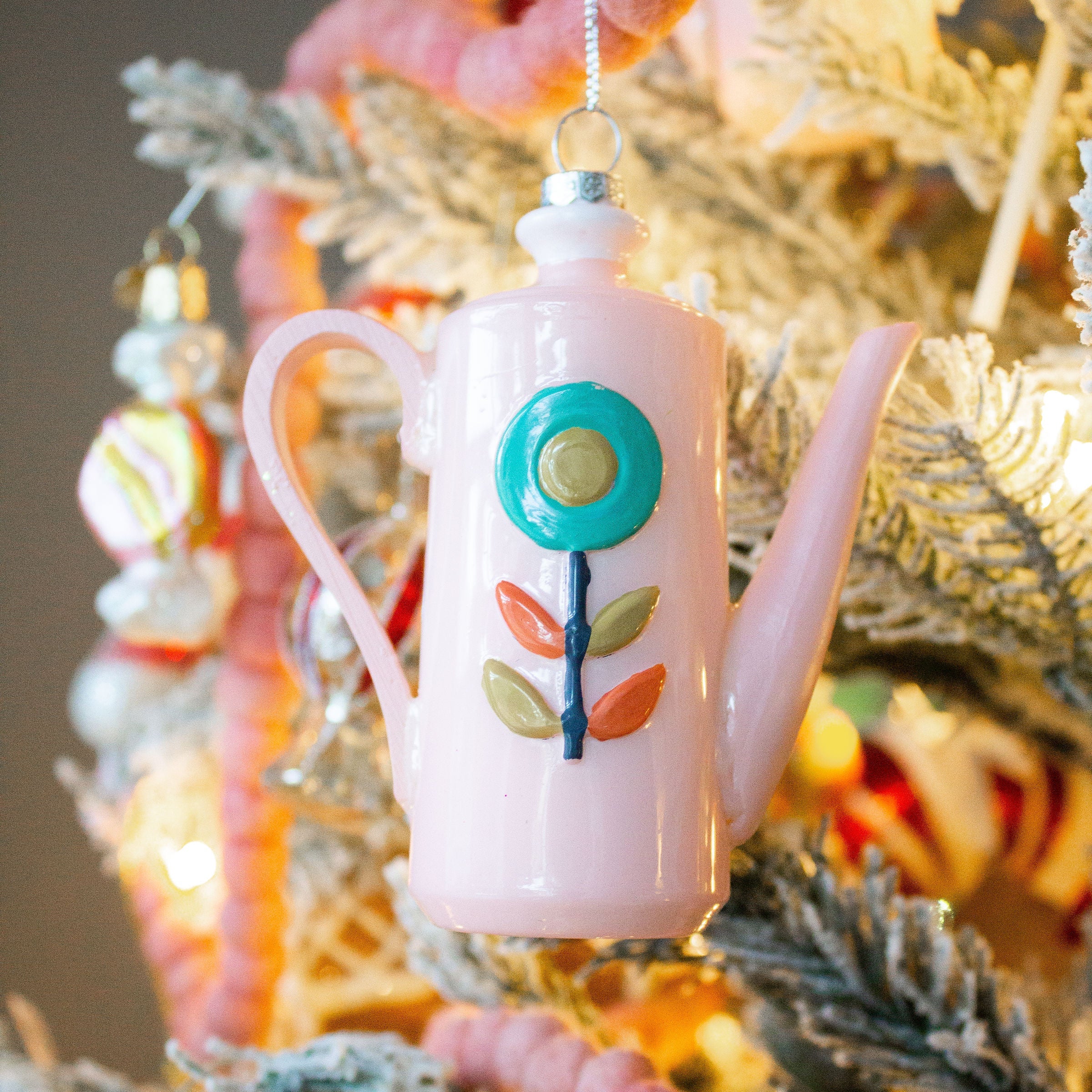 A vintage-style coffee pot ornament with a pink body and floral pattern, hanging on a Christmas tree.