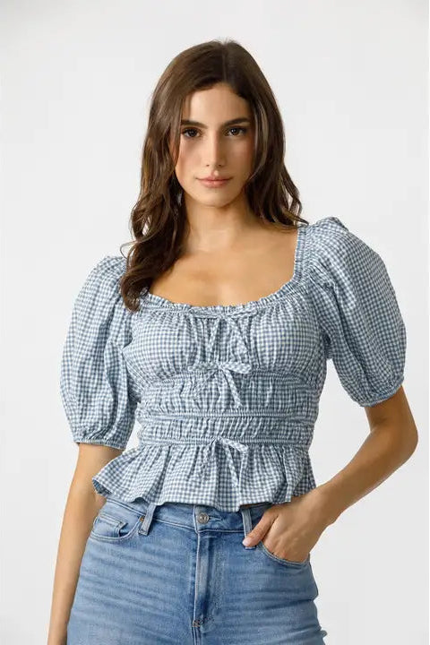 A woman wearing a blue and white checkered gingham top with a square neckline and puffed short sleeves, paired with blue jeans.