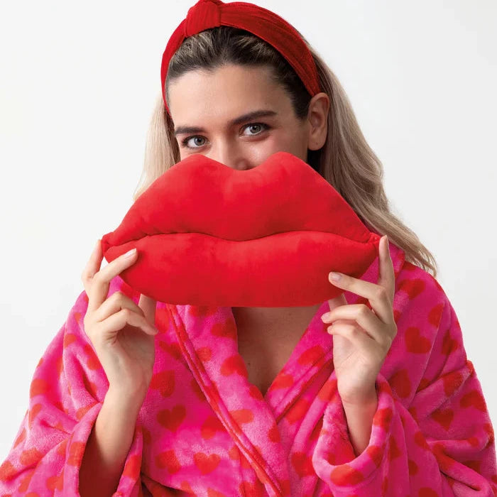 Woman in a pink robe holding a red lip-shaped pillow against her face on a white background