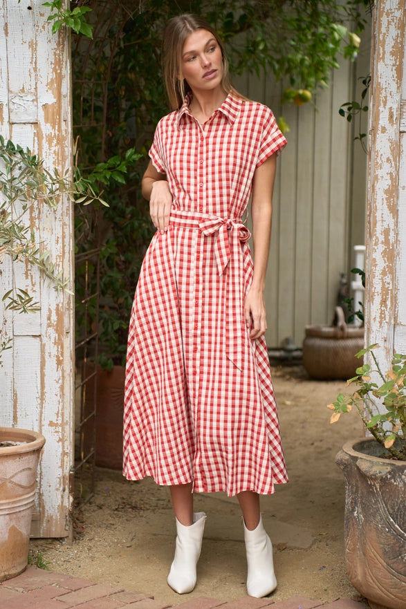 Woman wearing a red and white checkered dress standing in an outdoor setting with plants and rustic elements.