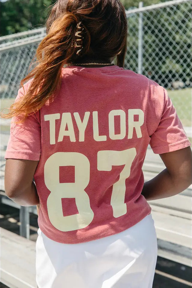 Person wearing a pink t-shirt with 'TAYLOR 87' printed on the back, standing outdoors.