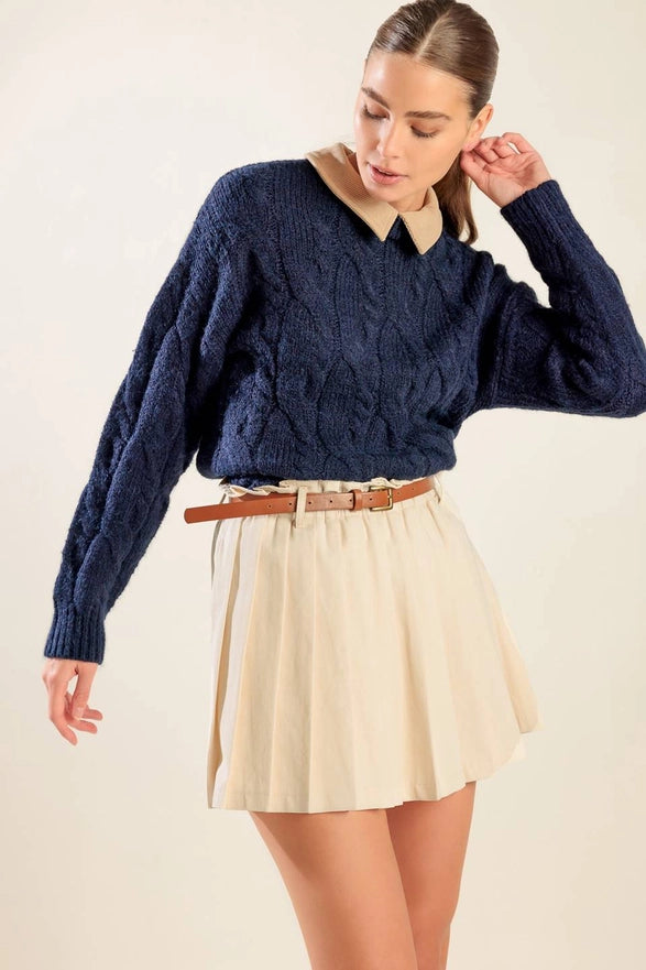 Woman wearing a blue cable knit sweater and beige pleated skirt on a plain background