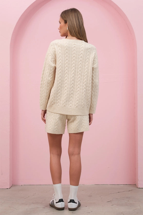 Woman wearing a cream cable knit sweater and shorts set against a pink archway.