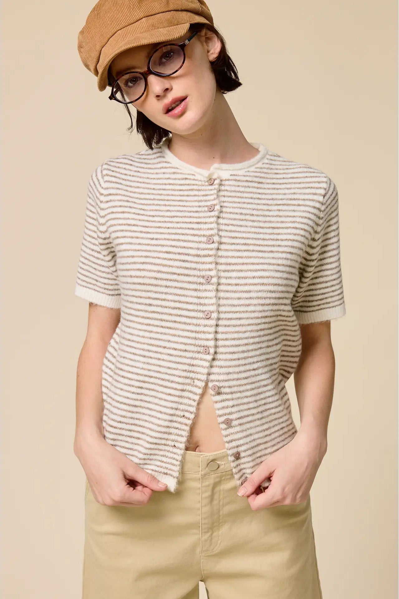 Person wearing a striped shirt, beige cap, and glasses against a beige background