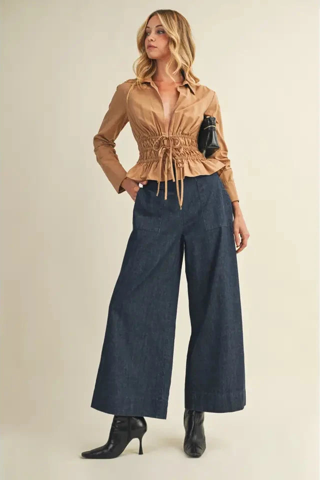 Woman wearing a tan blouse and blue wide-leg pants against a beige background