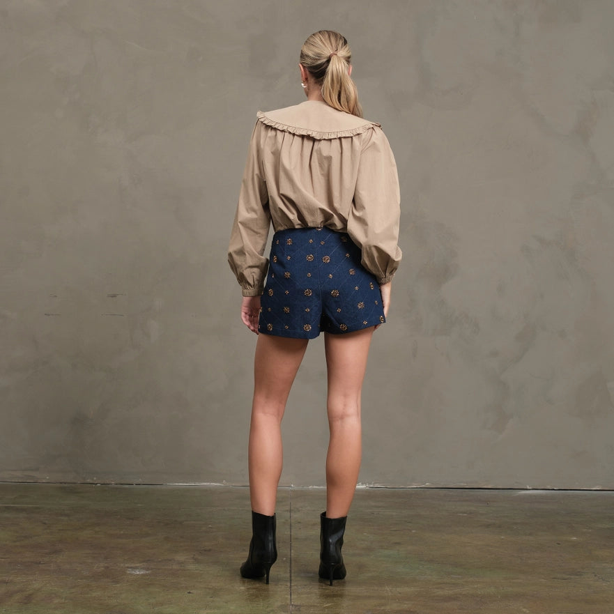 Woman wearing a beige jacket and blue patterned shorts against a gray wall.