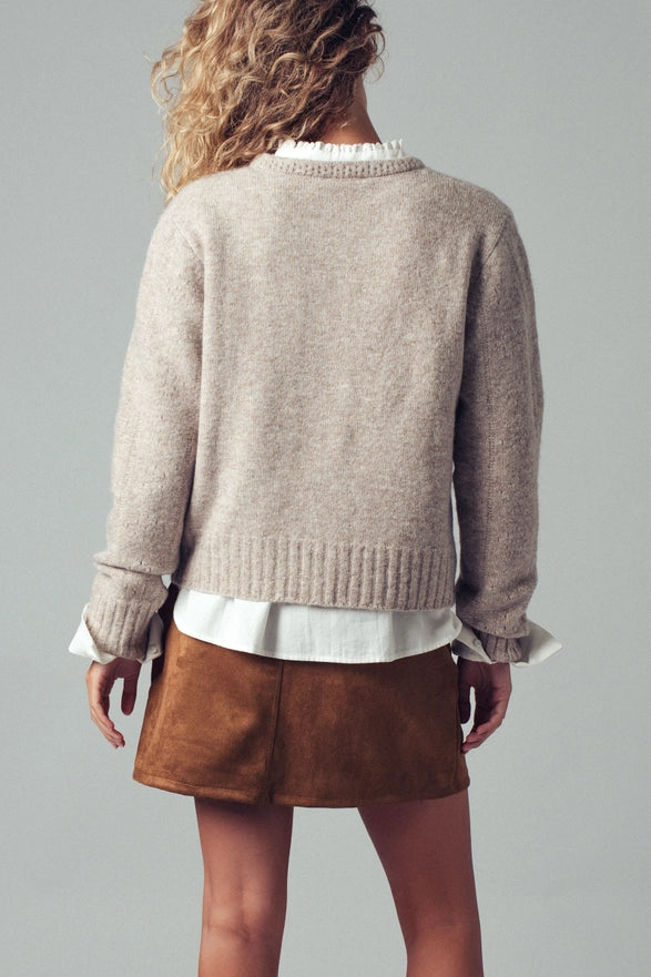 Person wearing a beige sweater over a white shirt with brown shorts on a gray background