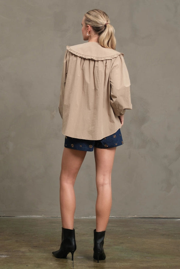 Woman wearing a beige jacket and dark shorts standing against a plain wall.