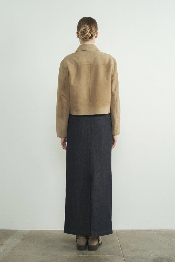 Person wearing a beige jacket and dark skirt standing against a white wall.