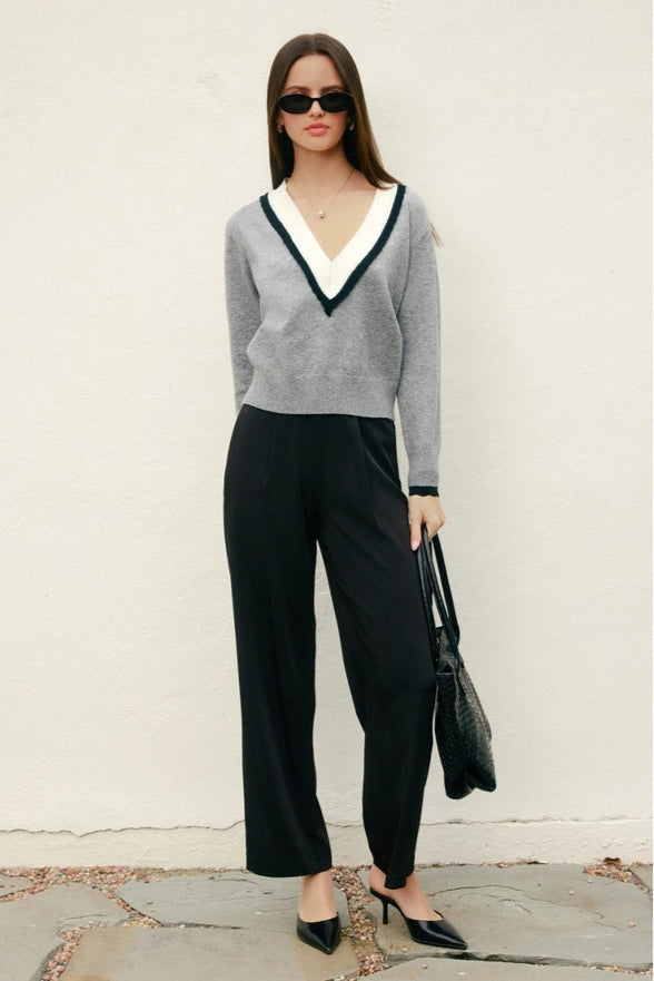 Woman wearing a gray sweater with a V-neck and black pants against a white wall.