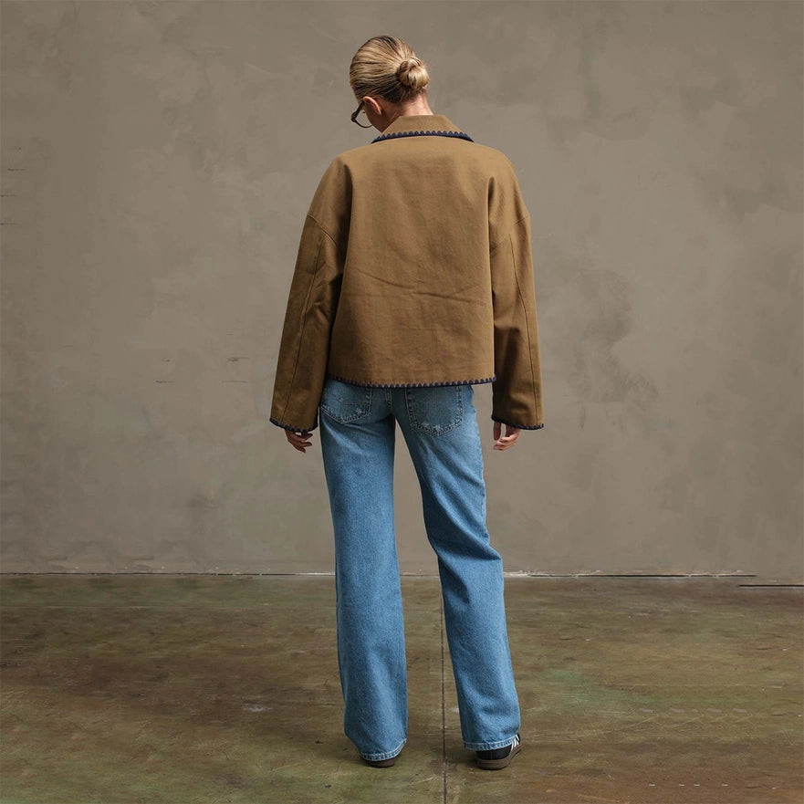 Person wearing a brown jacket and blue jeans standing against a plain wall.
