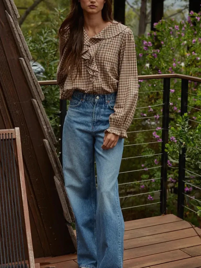 Woman wearing a plaid shirt and blue jeans standing on a wooden deck with greenery in the background