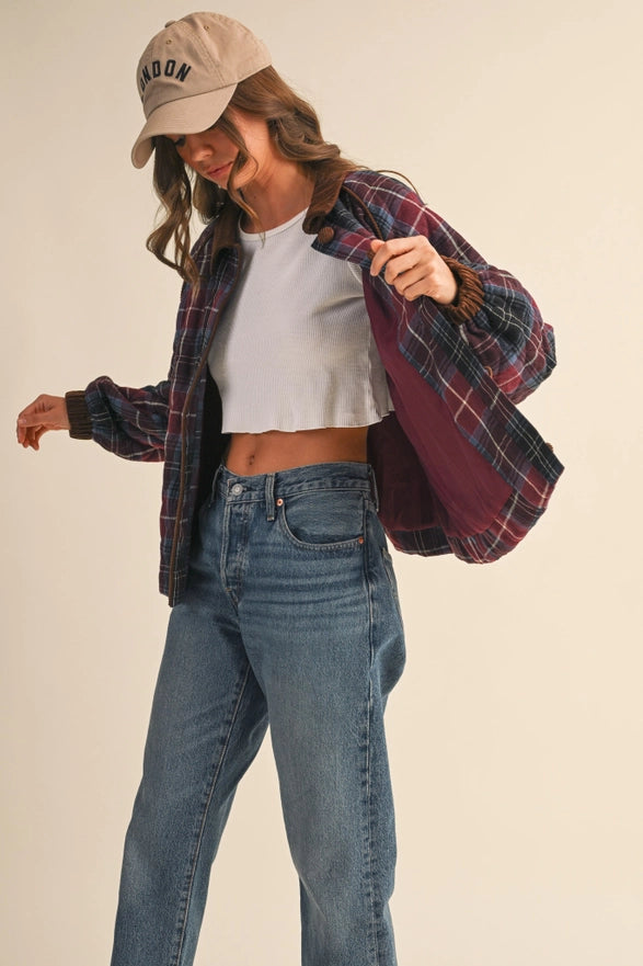 Person wearing a plaid shirt, white crop top, and blue jeans with a beige cap on a plain background