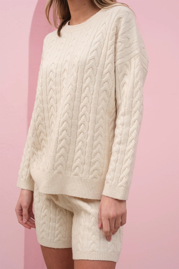Person wearing a cream cable knit sweater and shorts against a pink background