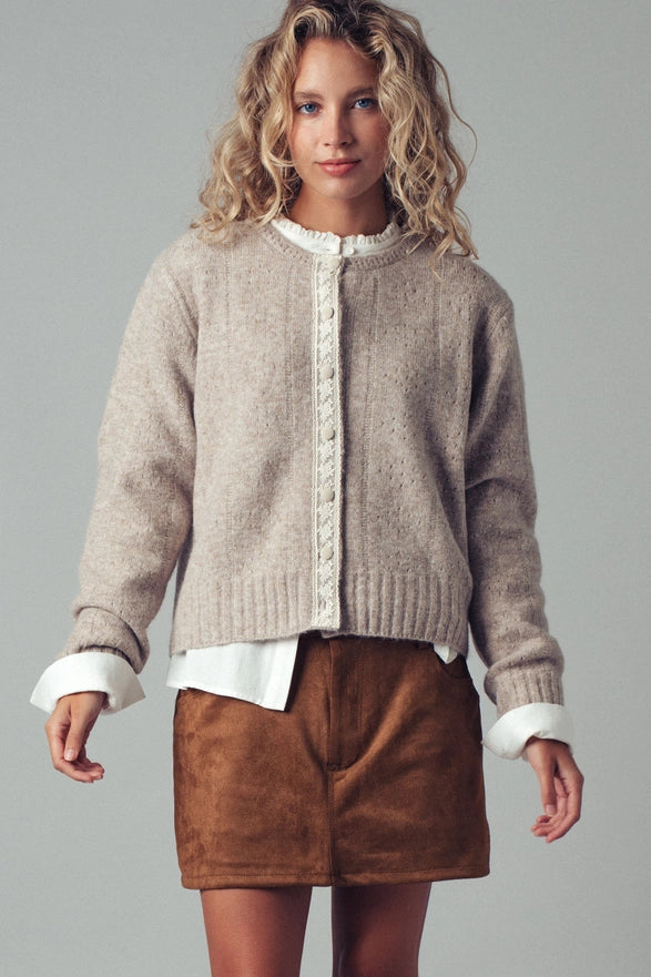Woman wearing a beige cardigan over a white shirt with brown shorts on a gray background