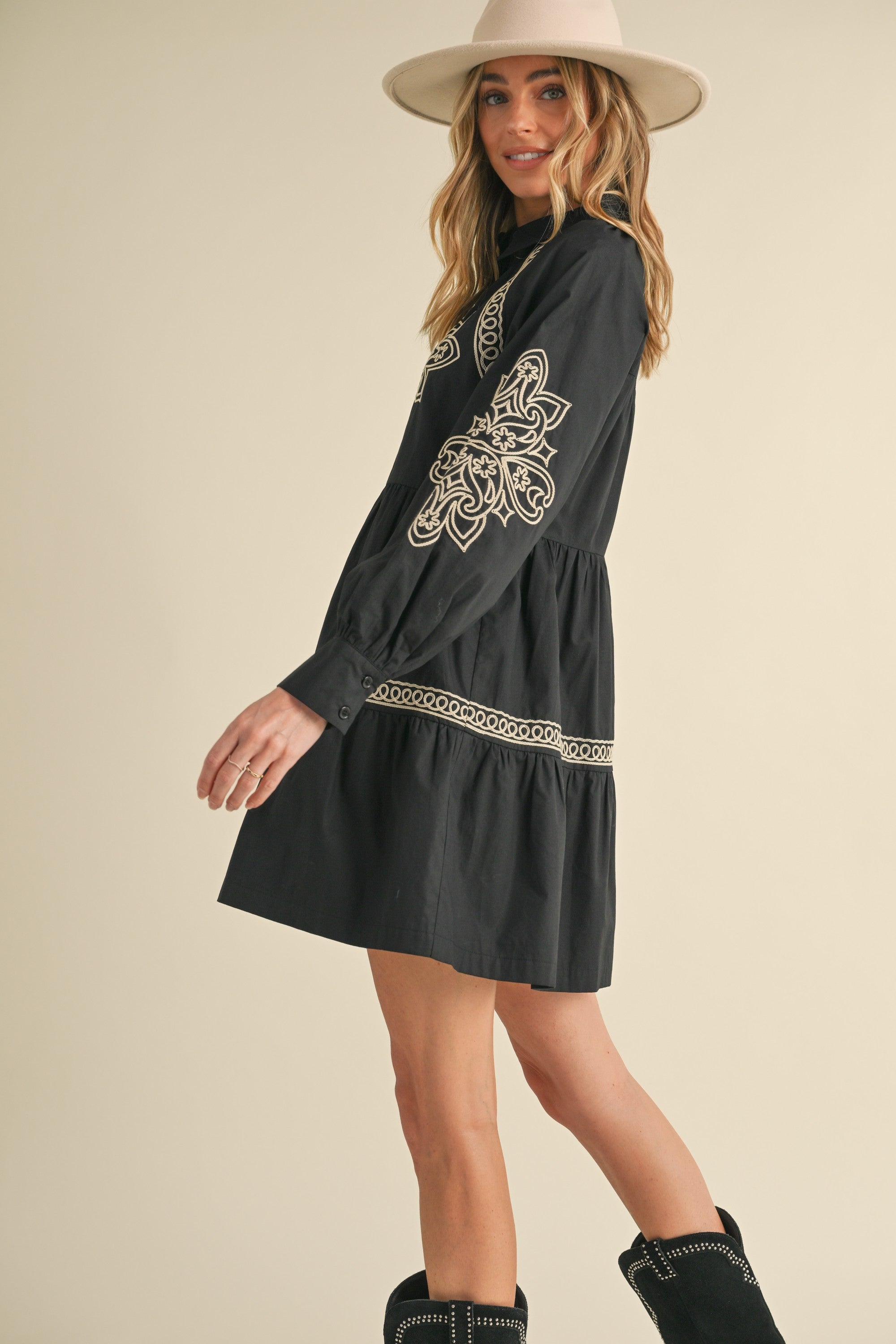 Woman wearing a black dress with white embroidery, black boots, and a beige hat on a beige background