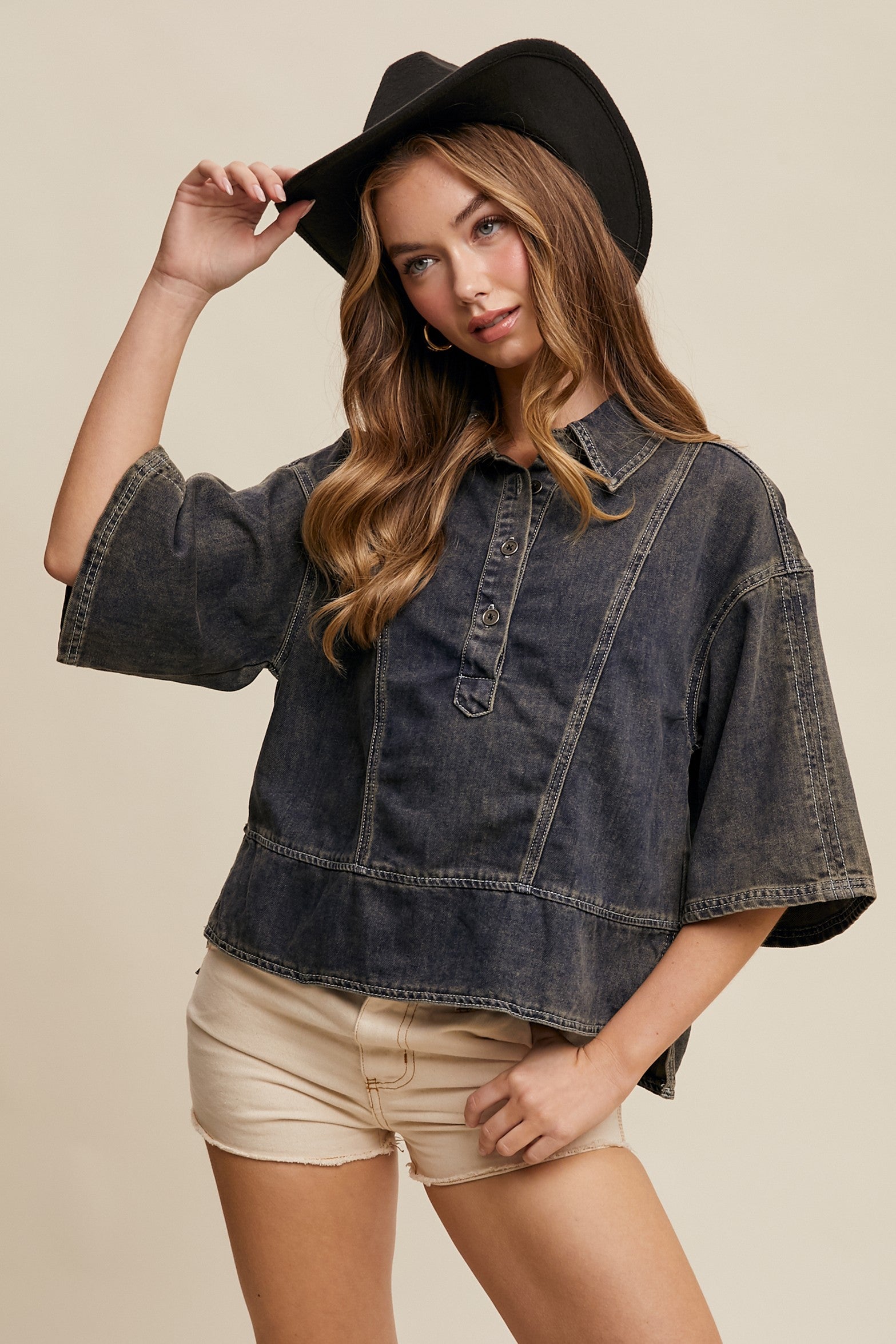 A female model wearing an oversized, collared, denim top with a partial button placket, in a washed denim finish. She is also wearing light-colored shorts and a wide-brimmed hat.
