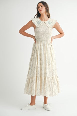 A woman wearing a floral printed sleeveless midi dress with a smocked bodice and embroidery on the collar. The dress has a ruffle detail on the collar and a button keyhole closure at the back neckline.