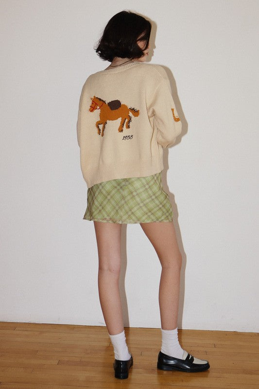 Person wearing a beige sweater with a horse design and a green plaid skirt against a white wall.