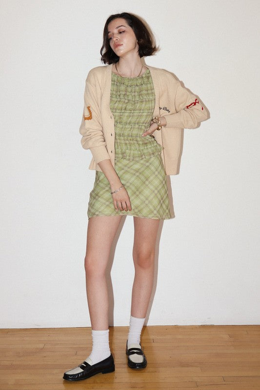 Woman wearing a green checkered dress and beige cardigan against a white wall.
