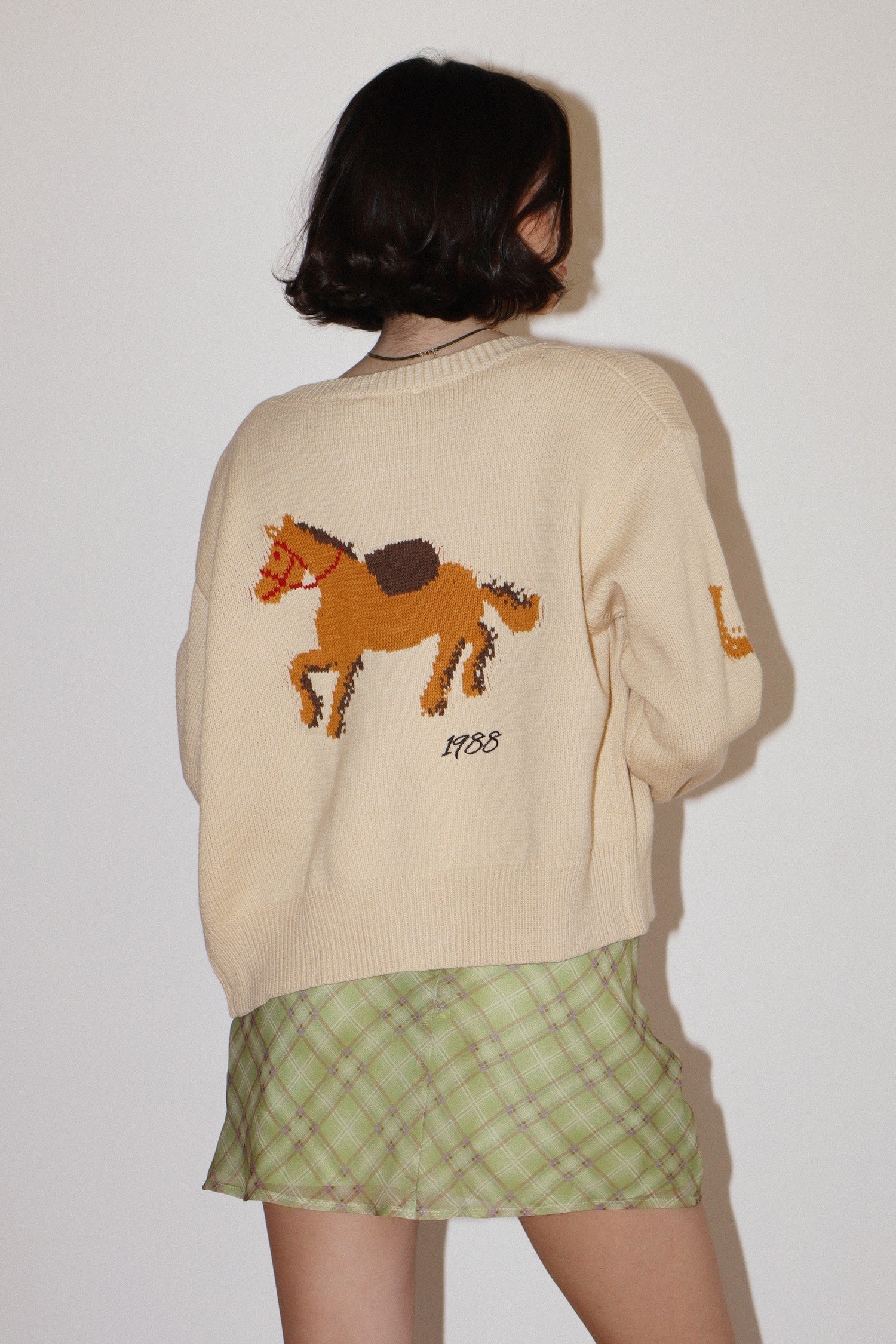 Person wearing a beige sweater with a horse design and a green plaid skirt on a plain background