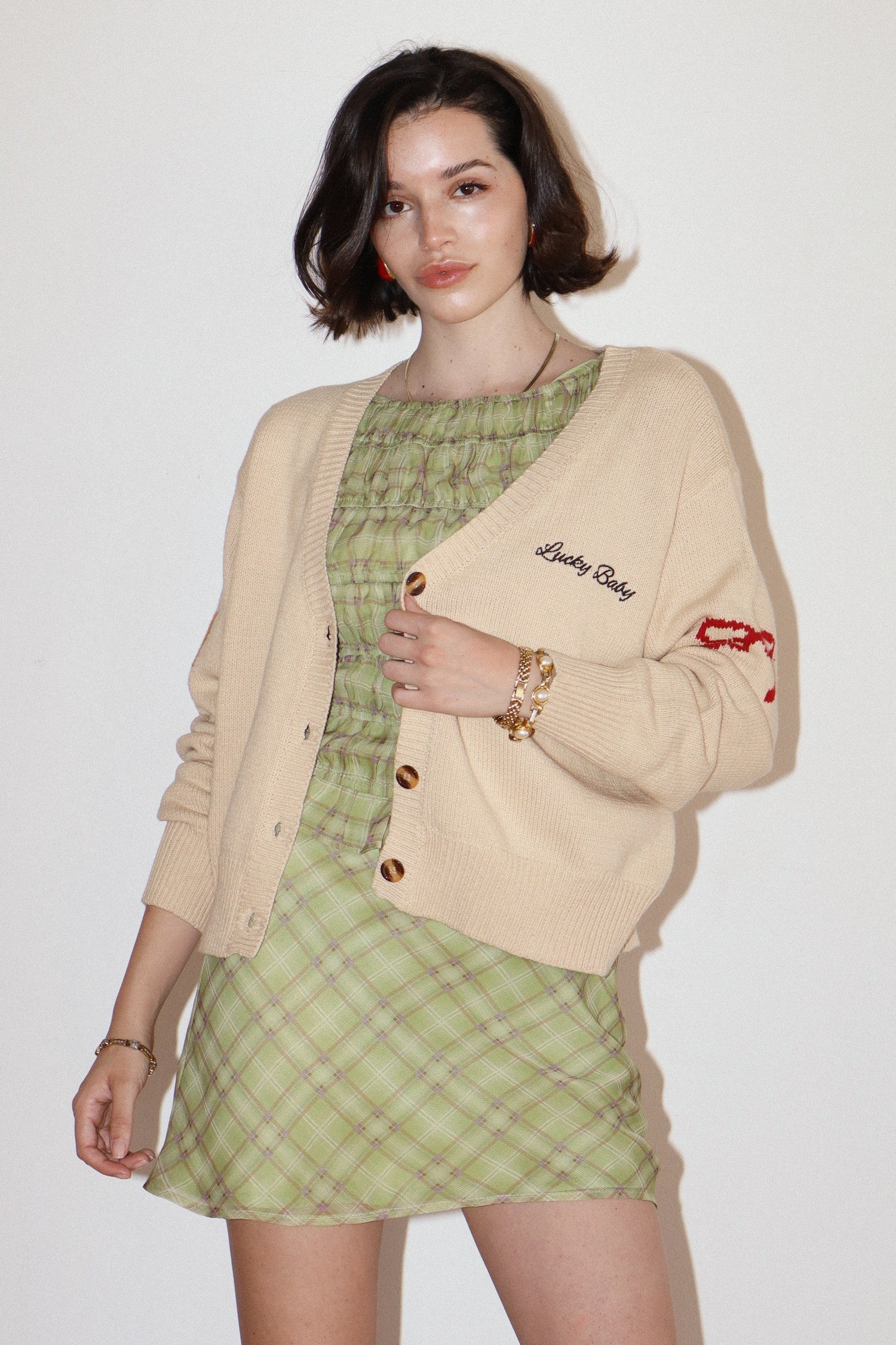 Woman wearing a beige cardigan with embroidered text and a green plaid dress on a white background