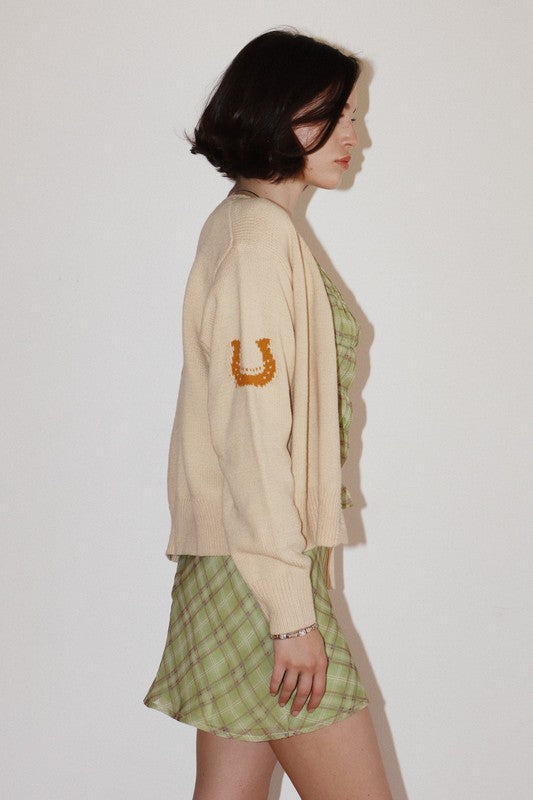 Woman wearing a beige cardigan with a horse shoe design and a green plaid dress on a white background