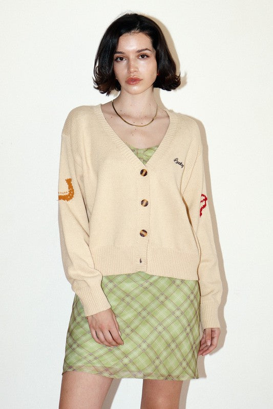 Woman wearing a beige cardigan with embroidered designs and a green plaid skirt on a white background
