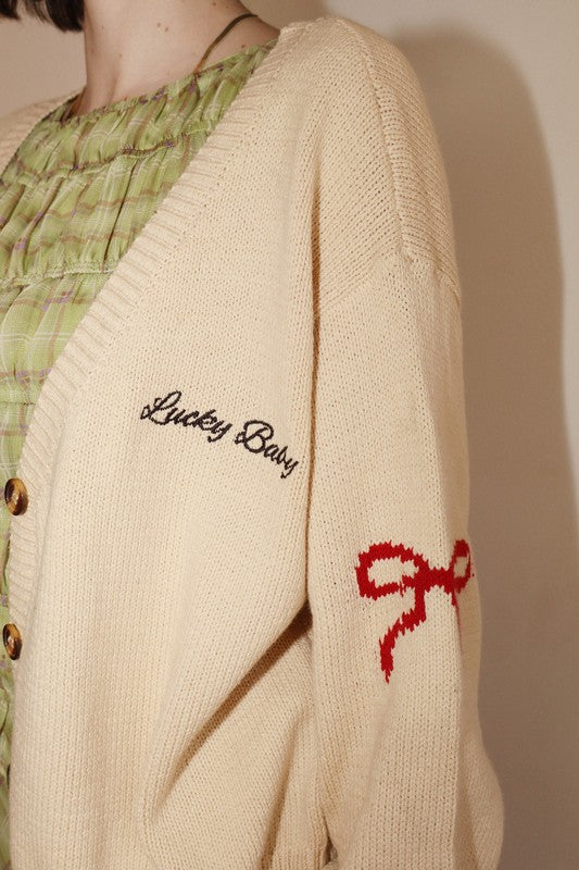 Beige cardigan with 'Lucky Baby' text and red bow design on a plain background