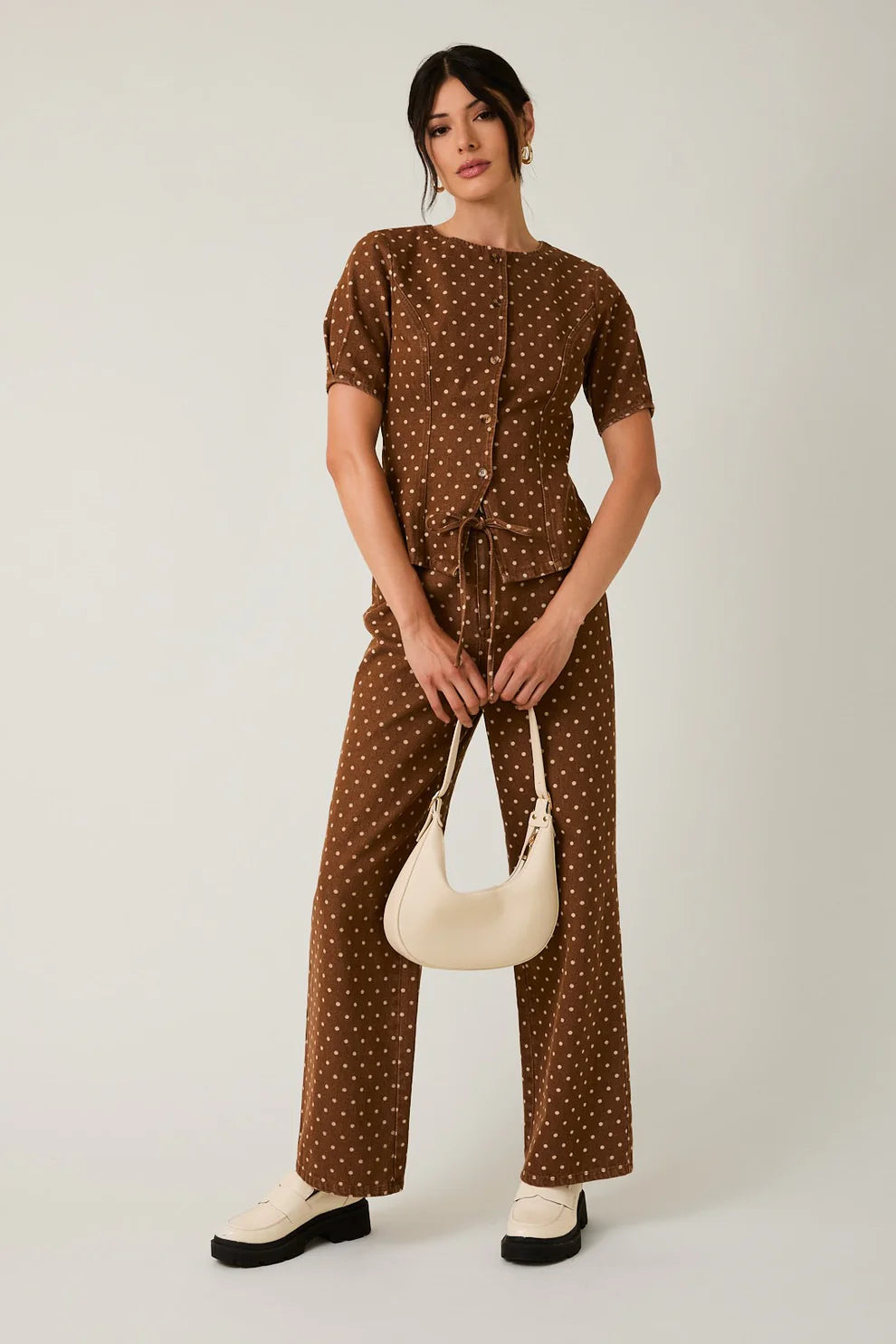 Woman wearing a brown polka dot jumpsuit holding a beige handbag on a white background