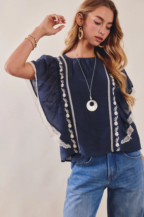 Woman wearing a navy top with white embroidery and blue jeans on a plain background