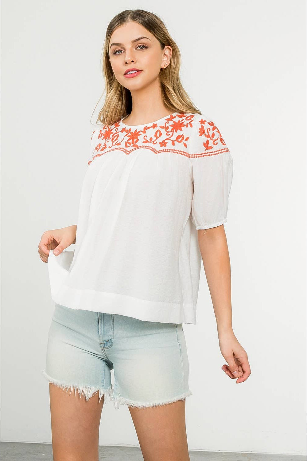 Woman wearing a white blouse with orange embroidery and light blue shorts on a white background