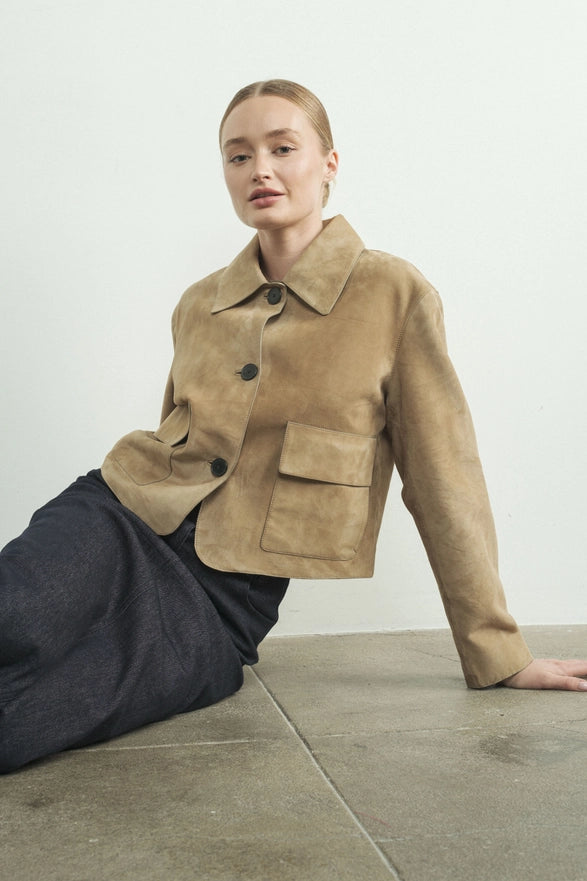 Person wearing a beige jacket sitting on a concrete floor with a white wall background