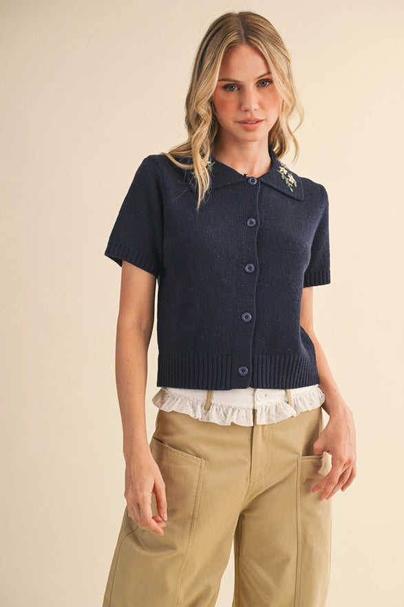 Woman wearing a navy short-sleeve cardigan with a white lace trim over a beige outfit on a beige background