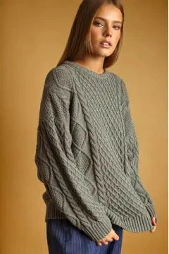 A woman wearing an oversized cable-knit sweater in a muted sage green color, paired with blue jeans, set against a solid warm tan background.