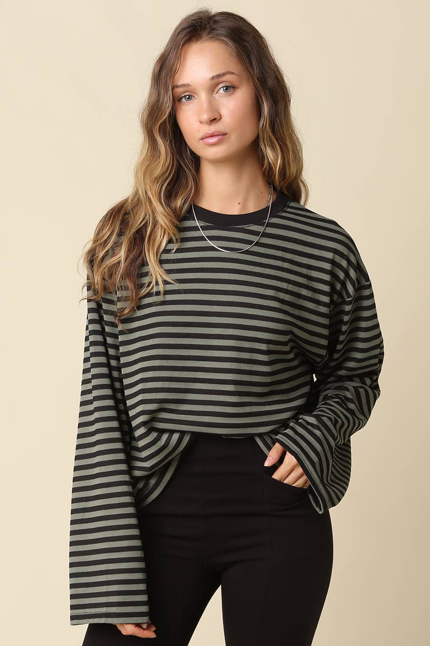 A woman modeling a long-sleeve striped top with black bell sleeves, featuring horizontal stripes in dark green and black, paired with black pants against a solid beige background.